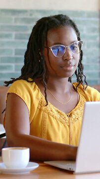 Woman working on laptop in cafe, focusing then removing glasses, inserting earbuds for remote work