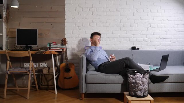 Man in a shirt and pants is lying on the sofa and tossing crumpled paper toward a trash can, frustrated by creative block while working remotely in his home office