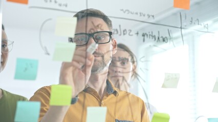 Business colleagues brainstorming, writing strategy notes on glass board, collaborating with colorful sticky notes in creative workplace meeting © volha_r