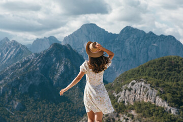 Fototapeta premium Rear view of a young female tourist in a summer dress standing on a cliff edge. She is holding her hat against the wind while admiring the breathtaking panoramic view of the rocky mountain landscape.