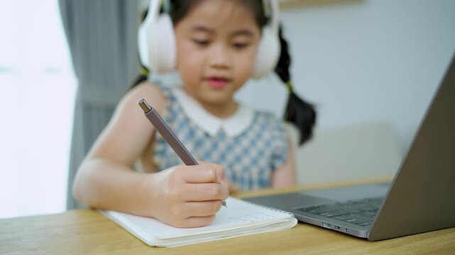 Young Girl in Headphones Writing Notes at Desk with Laptop in Bright Room, Engaged in Creative Learning Activity, Focused on Education