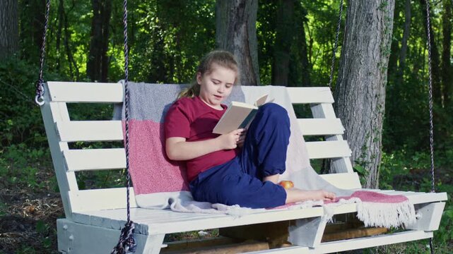 Kid studies happily on a swing in summertime. Reading book helps child develop knowledge. Boy sits on swing and reads with interest. Child enjoys literacy and learning outdoors.