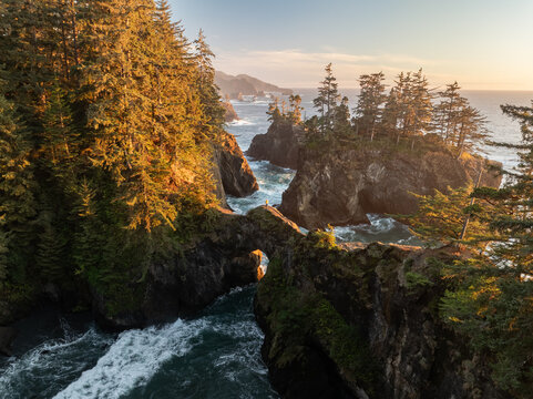 Aerial view of the Natural Bridges rock formations along the rugged coastline with dense evergreen forests and crashing waves during golden hour in Brookings, Oregon, United States.