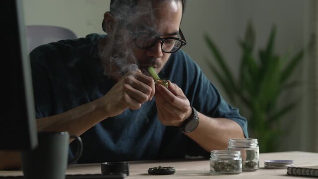 Man smoking a cannabis from smoking pipe, with cannabis equipment and accessories on the table.
