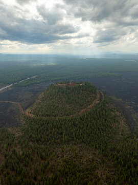 Aerial view of Lava Butte cinder cone covered in evergreen trees under a dramatic cloudy sky with sun rays in Deschutes County, Oregon, United States.