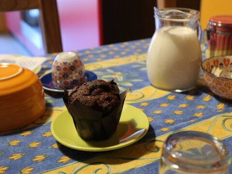 Muffin al cioccolato servito su un piattino verde acido accanto a una brocca di vetro piena di latte fresco su una tavola da colazione colorata e allegra.