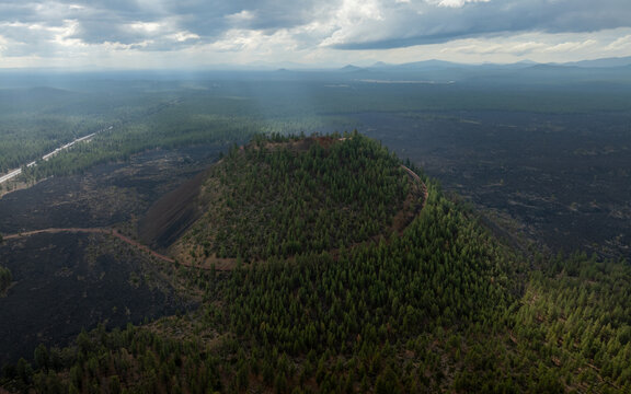 Aerial view of Lava Butte cinder cone covered in pine trees surrounded by dark volcanic rock and vast evergreen forests under a dramatic cloudy sky Deschutes County, Oregon, United States.