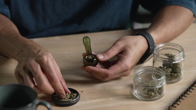 Man filling glass pipe with cannabis, with cannabis equipment and accessories on the table.