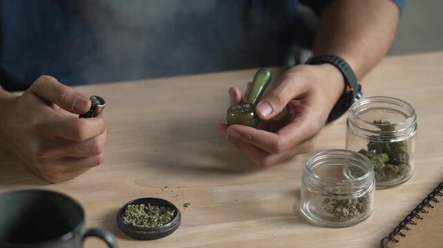 Man smoking a cannabis from smoking pipe, with cannabis equipment and accessories on the table.