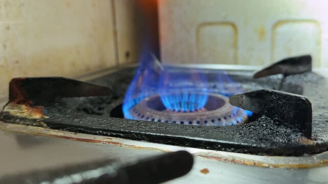 a gas stove burner turned on with blue flame, rusted grate, and stained kitchen tiles, showing active heat for cooking in an indoor kitchen setting.