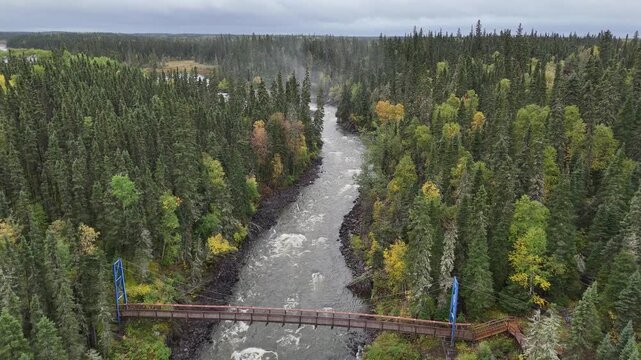 Aerial drone flies high above the Grass River in northern Manitoba, Canada, following it upstream toward a swinging bridge. Rapids under the bridge and river meanders visible with fall foliage.