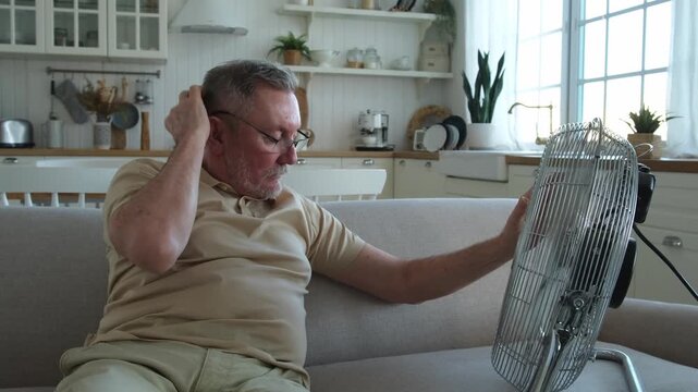 Senior man refreshing himself near fan during summer heat wave hot day. Elderly man suffering from summer heat trying to survive with electric fan. Man enjoying refreshing air blowing from ventilator