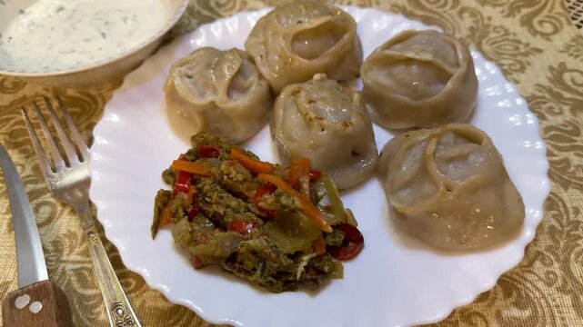 Large Manti dumplings on white plate with herbed yogurt sauce in bowl, spiced vegetable salad, fork and knife resting on patterned