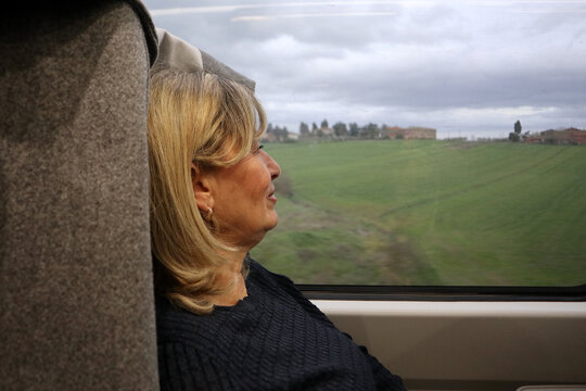 Donna bionda di profilo seduta sul treno che guarda fuori dal finestrino il paesaggio di campagna scorrere durante un viaggio