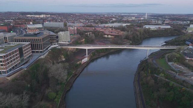 High-angle aerial drone shot flying backward from the modern Keel Crossing to reveal the historic Monkwearmouth Railway Bridge and Wearmouth Bridge at dawn.