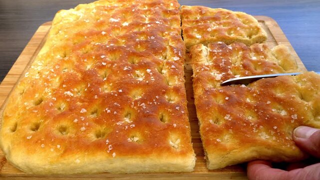 Closeup of scissors cutting a hand held batch of warm, oven baked, focaccia Italian bread, a classic recipe of flat, dimpled dough topped with rosemary, olive oil and sea salt flakes.