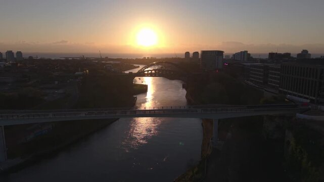 Wide aerial drone shot tracking forward toward the Keel Crossing at sunrise, with the silhouetted Wearmouth Bridge and Monkwearmouth Railway Bridge spanning the River Wear in the background.