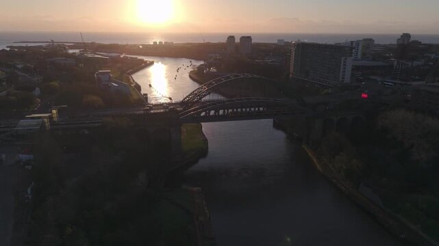 Wide aerial drone shot tracking slowly toward the silhouetted Wearmouth Bridge and Monkwearmouth Railway Bridge at sunrise, featuring a Tyne and Wear Metro train crossing the River Wear.