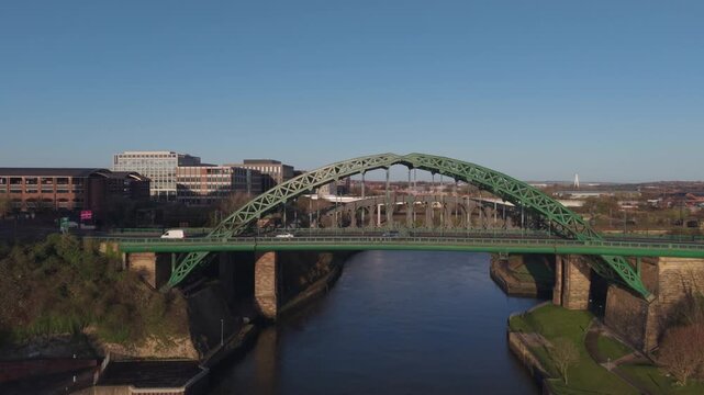 Wide aerial drone shot tracking slowly toward the Wearmouth Bridge and Monkwearmouth Railway Bridge in Sunderland during the morning commute, showing city traffic and riverside architecture.