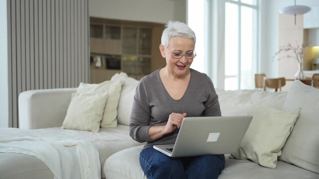 Confident stylish happy middle aged senior woman using laptop at home. Stylish older mature 60s grandmother sitting at couch looking at computer screen typing chatting reading writing email