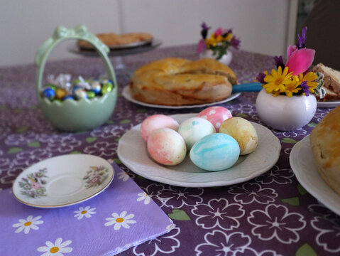 Tavola imbandita per la colazione di Pasqua con uova sode colorate, pane dolce tradizionale, ovetti di cioccolato e decorazioni floreali su tovaglia lilla a fiori