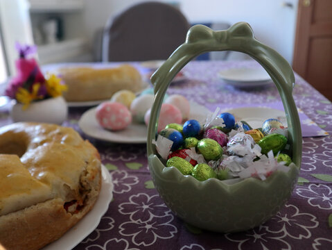 Tavola imbandita per la colazione di Pasqua con uova sode colorate, pane dolce tradizionale, ovetti di cioccolato e decorazioni floreali su tovaglia lilla a fiori