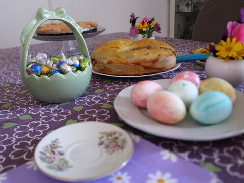 Tavola imbandita per la colazione di Pasqua con uova sode colorate, pane dolce tradizionale, ovetti di cioccolato e decorazioni floreali su tovaglia lilla a fiori