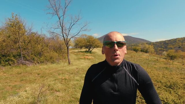 Active mature man in his 50s running on a trail through a scenic field and forest on a sunny spring day. Healthy senior lifestyle, jogging and exercising outdoors. High quality 4k footage.