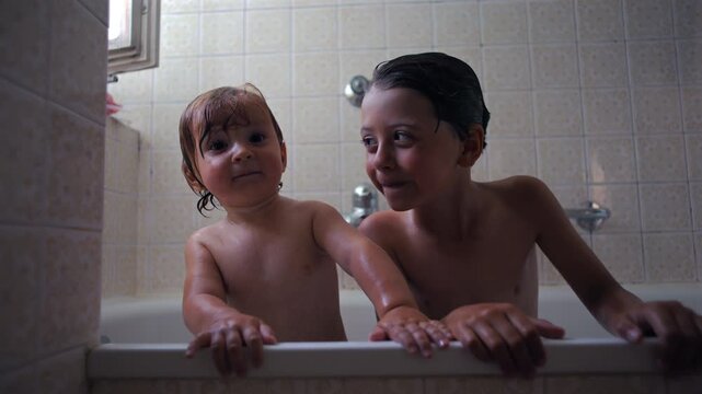 Baby and older sibling leaning on bathtub edge during bath time, curious expressions, playful water moment, family routine, hygiene and bonding in home bathroom environment