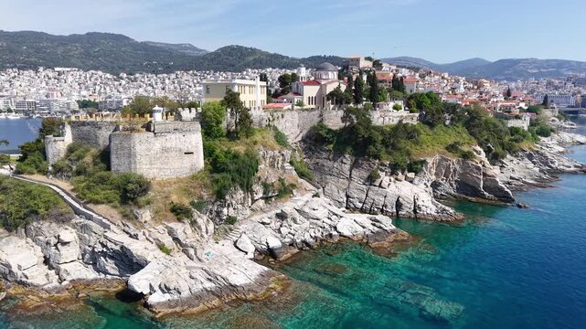 Aerial shot of a historic-looking coastal town on a rocky peninsula with fortified walls and turquoise sea.