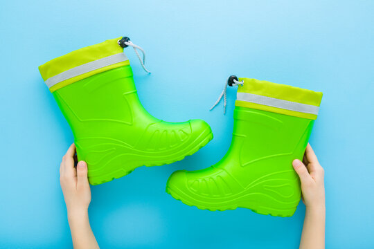 Little child boy hands holding and showing new clean green rubber boots on light blue table background. Pastel color. Closeup. Point of view shot. Flat lay. Top down view.
