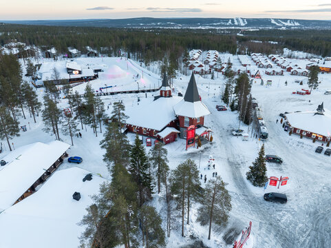 Aerial view of Santa Claus Village with snow-covered wooden cabins and pine trees under a soft winter sky in Rovaniemi, Lapland, Finland.