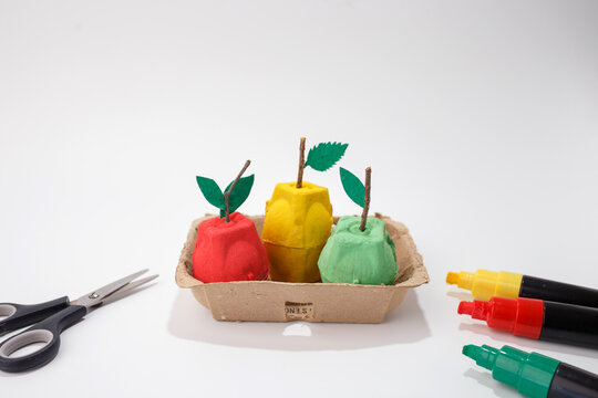 Three painted apple shapes made from egg carton with paper leaves in cardboard tray with scissors and markers on white background. Recycling and kids education concept. Eye-level composition 
