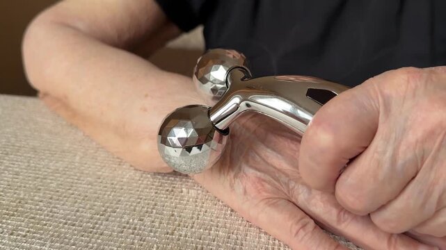 Elderly woman massaging wrinkled arm with silver dual roller massager on textured fabric surface, slow gentle strokes, focus.