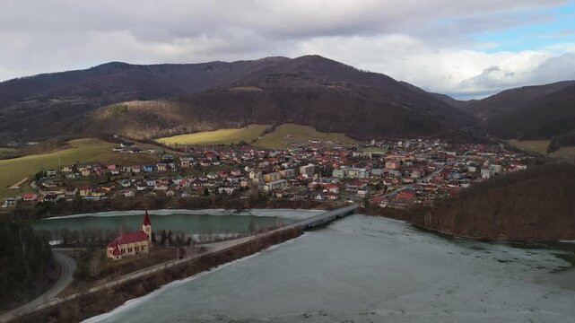 Margecany village in eastern Slovakia features the Kostol sv Margity Catholic church surrounded by hills, forests, and the frozen Ruž&iacute;n reservoir under an overcast winter sky in this aerial panorama.