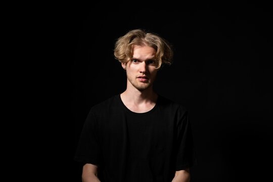 Young man with light stubble and blonde curly hair, wearing a black t-shirt, standing against a dark background, looking with a confused and intense expression directly at the viewer