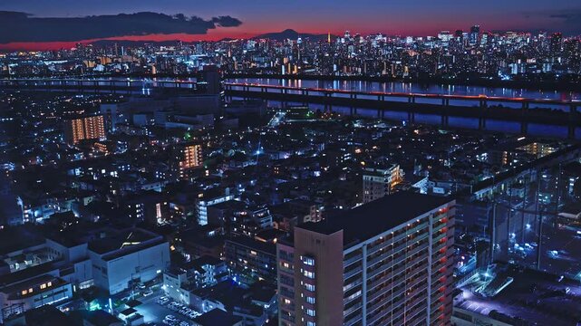 A high-angle drone shot capturing the sprawling urban landscape of Tokyo as night falls. Twinkling lights illuminate the city and river under a deep blue and crimson twilight sky.