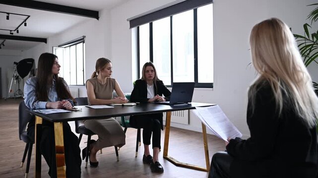 Young woman job candidate sits across three hr managers at a modern office table, answering questions and holding her resume during a professional interview and recruitment meeting