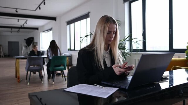 Professional woman with blonde hair sitting at her desk in a contemporary open plan office, using her smartphone for messaging while working on a laptop, with colleagues in the background