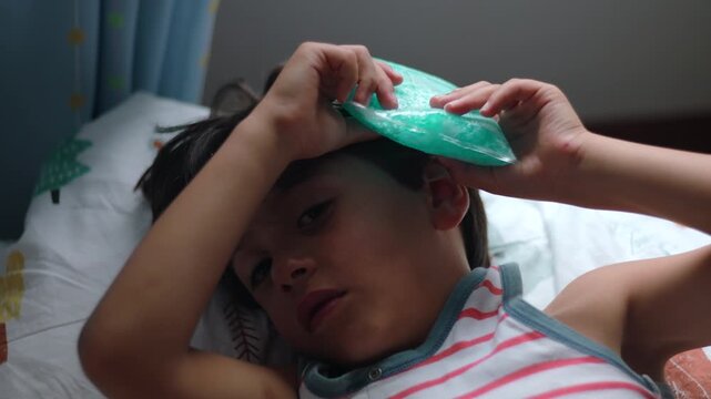 Close view of child adjusting cold pack on head while lying down showing discomfort fatigue and attempt to soothe pain during recovery in home environment
