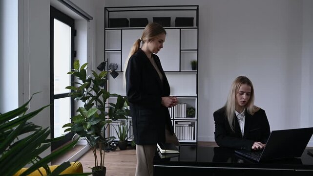 Young female manager scolding her subordinate for a work mistake while the employee stands listening with a guilty expression and the boss works on her laptop in a modern office interior