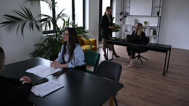 Young female candidate confidently answering questions during a job interview with a human resources manager in a modern office, while other employees are working in the background