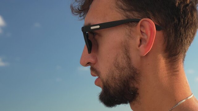 Man bearded sunglasses, young male looking away then turning to camera under clear blue sky