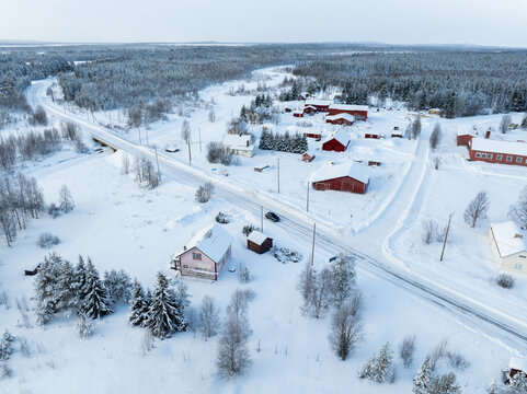 Aerial view of traditional red wooden houses and a snow-covered road crossing a bridge over a frozen river in a winter forest landscape in Lapland, Finland.