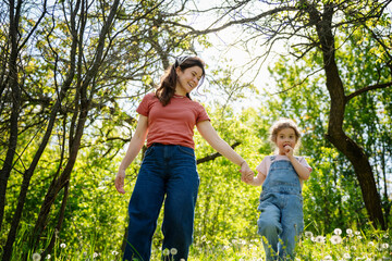 Fototapeta premium Happy mother and little daughter holding hands and walking together in sunny summer garden