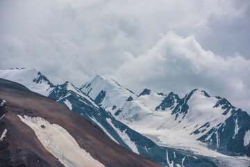 Dramatic view to large rock mountain range with snow-capped pinnacle in gray cloudy sky. Atmospheric landscape with snow cornice on snow-white pointy peak. Big rocky mountains and snowy peaked top. © Daniil