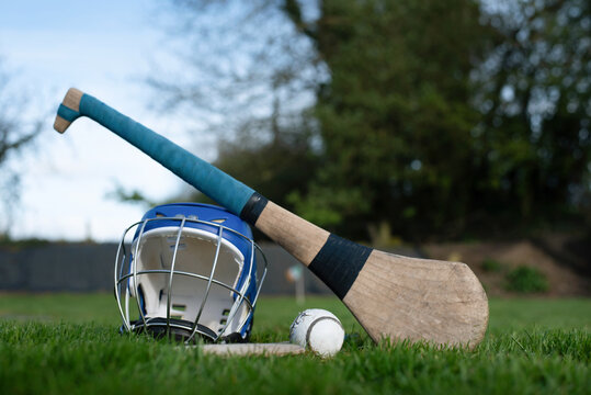 Hurling equipment arranged on a grassy pitch: a wooden hurley, sliotar and protective helmet with cage, evoking Gaelic sport, teamwork and outdoor play on rural green field