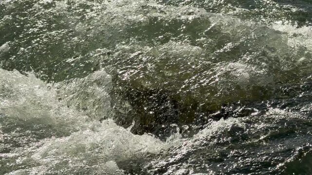 Fast river water crashes and foams over hidden stones. The bright sun makes the splashing waves sparkle, creating an energetic and powerful feeling of nature in motion.
