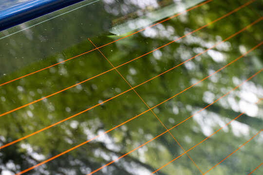 Close up of orange heating elements on a car rear window glass with reflections of green trees in daylight
