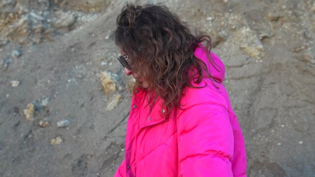 Woman fashion jacket outdoors standing by a sandy slope in a bright pink puffer coat while preparing to change outfits